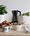 Caffettiera Elettrica DeMaison nera 6 tazze in ambiente colazione con tazza di caffè, croissant e cereali, moka elettrica elegante per una perfetta prima colazione quotidiana
